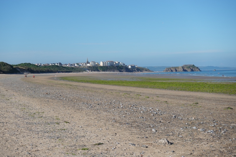 Der weite Strand bei Penally mit dem Blick auf Tenby im Hintergrund. Es ist Ebbe und das napoleonische Felsenfort ist mit der Küste verbunden. Tenby liegt erhöht auf den Klippen. Der Strand ist leer trotz des Sonnenscheins.