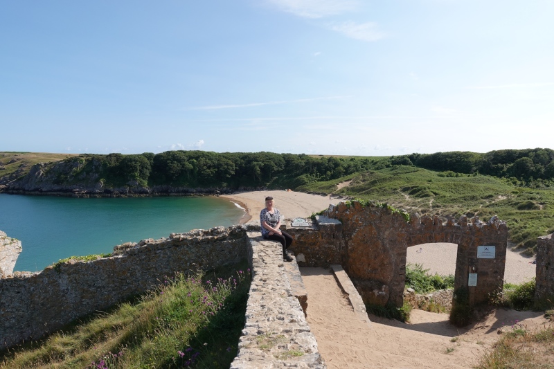 Die Autorin auf einer Mauer vor dem Strand Barafundle Bay Beach. Durch feinen Sand geht es hinab in die Bucht mit glasklarem Wasser, eingesäumt in Dünengras und von grünen Klippen eingeschlossen.