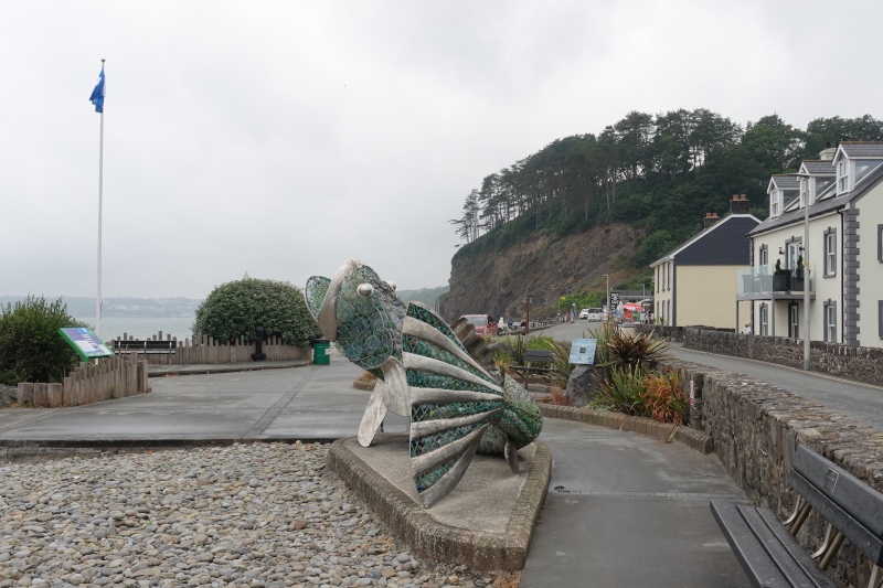 Ein kleiner Küstenort: im Vordergrund die Skulptur eines Fisches, der aus Plastikmüll gefertigt wurde. Eine kleine Straße führt am Strand entlang. Am Ende der Dorfstraße erheben sich hohe, bewaldete Klippen.