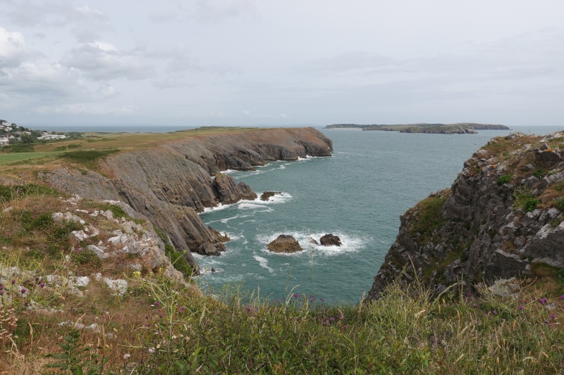 Ein weiter Blick über die Küste auf Caldey Island. Es ist bewölkt, die Wellen brechen sich an vorgelagerten Felsen.