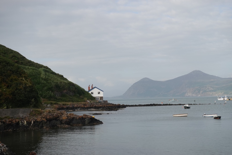 Eine Bucht mit natürlichem Hafen. Ein Fischerhaus steht am Rand der Klippen. Im Hintergrund erhebt sich das Yr Eifl Gebirge. Die See ist ruhig, der Himmel leicht bewölkt.