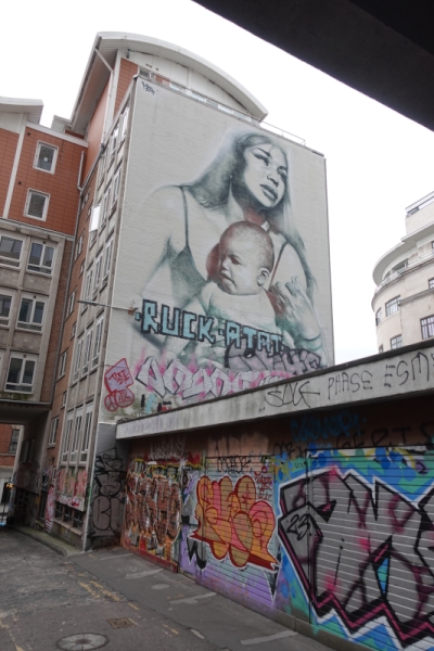 Auf der Seitenwand eines Wohnhauses das Bild einer jungen Mutter mit ihrem Baby. Beide schauen nicht wirklich glücklich. Im Vordergrund Garagen, die komplett mit Graffitis bemalt sind.