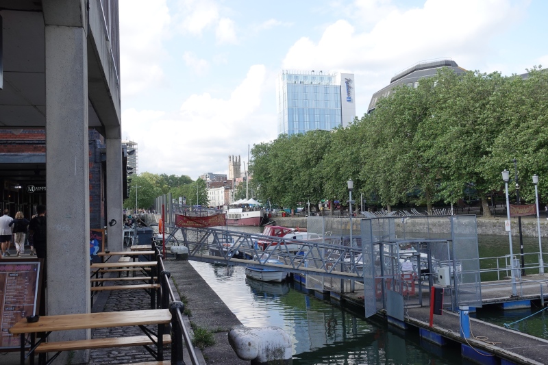 Man sieht einen Seitenarm des Flusses Avon in der Innenstadt von Bristol, an dessen Ufer sich verschiedene Pubs und Restaurants niedergelassen haben. Eine Reihe Kleiner Boote liegt hier vor Anker. Auf der gegenüberliegenden Seite führt ein alleeartiger Fussweg zum Fluss. Im Hintergrund sieht man den Tower des Radisson Hotels und den Eingang in die Altstadt.